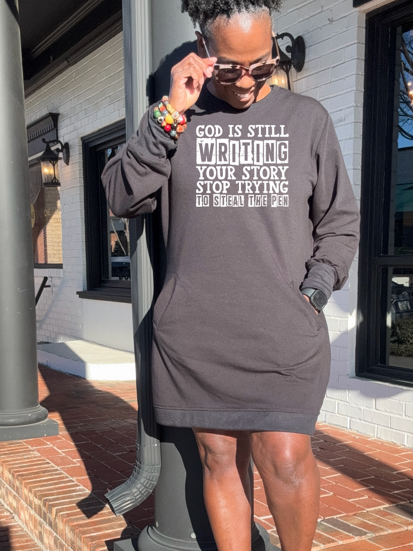 A black woman wearing a black sweatshirt dress that says God is Still writing your story standing outside an office