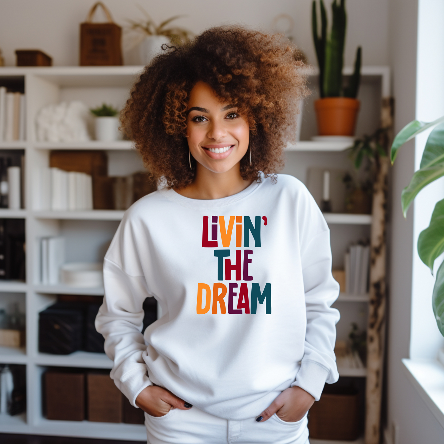 A black woman wearing a shite Livin The Dream sweatshirt, standing in her library