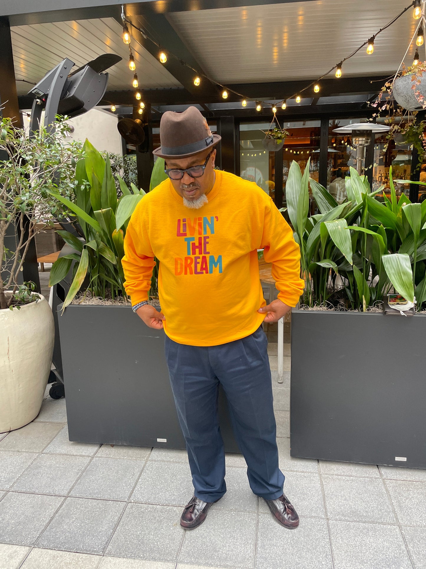 A Black man wearing a gold sweatshirt, that says Livin the dream. He is standing in an indoor setting with plants and furniture.