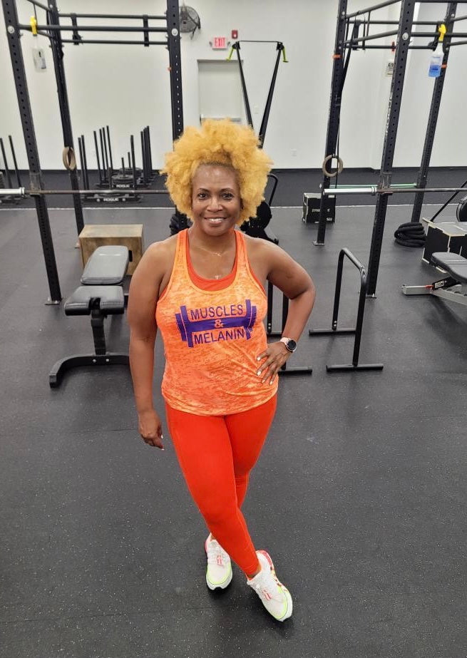 Black girls standing in a gyme wearing an orange muscles and melanin tank top
