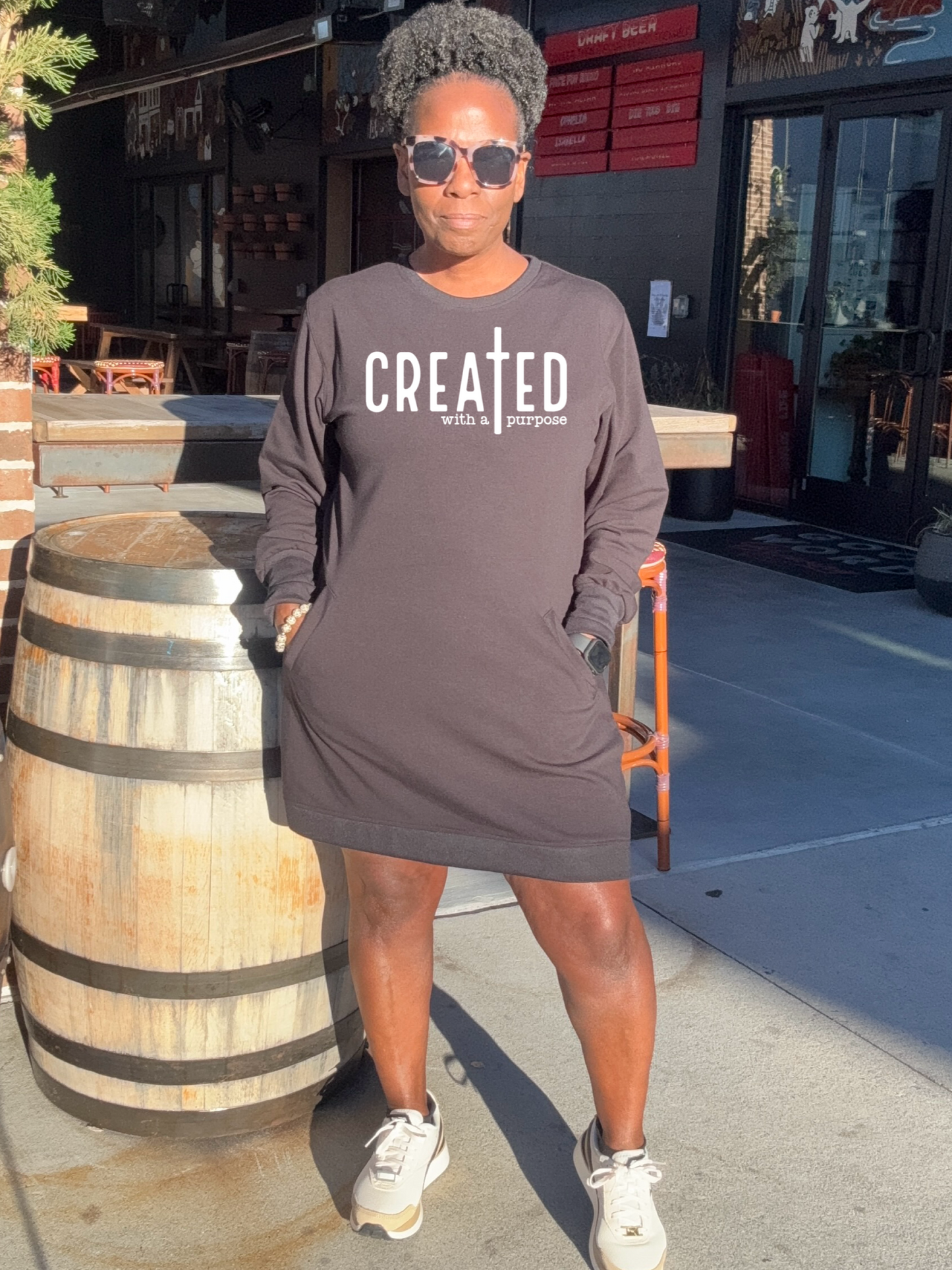 A Black woman standing outside a restaurant wearing a black sweatshirt dress with the text 'CREATED With A Purpose' on the front.