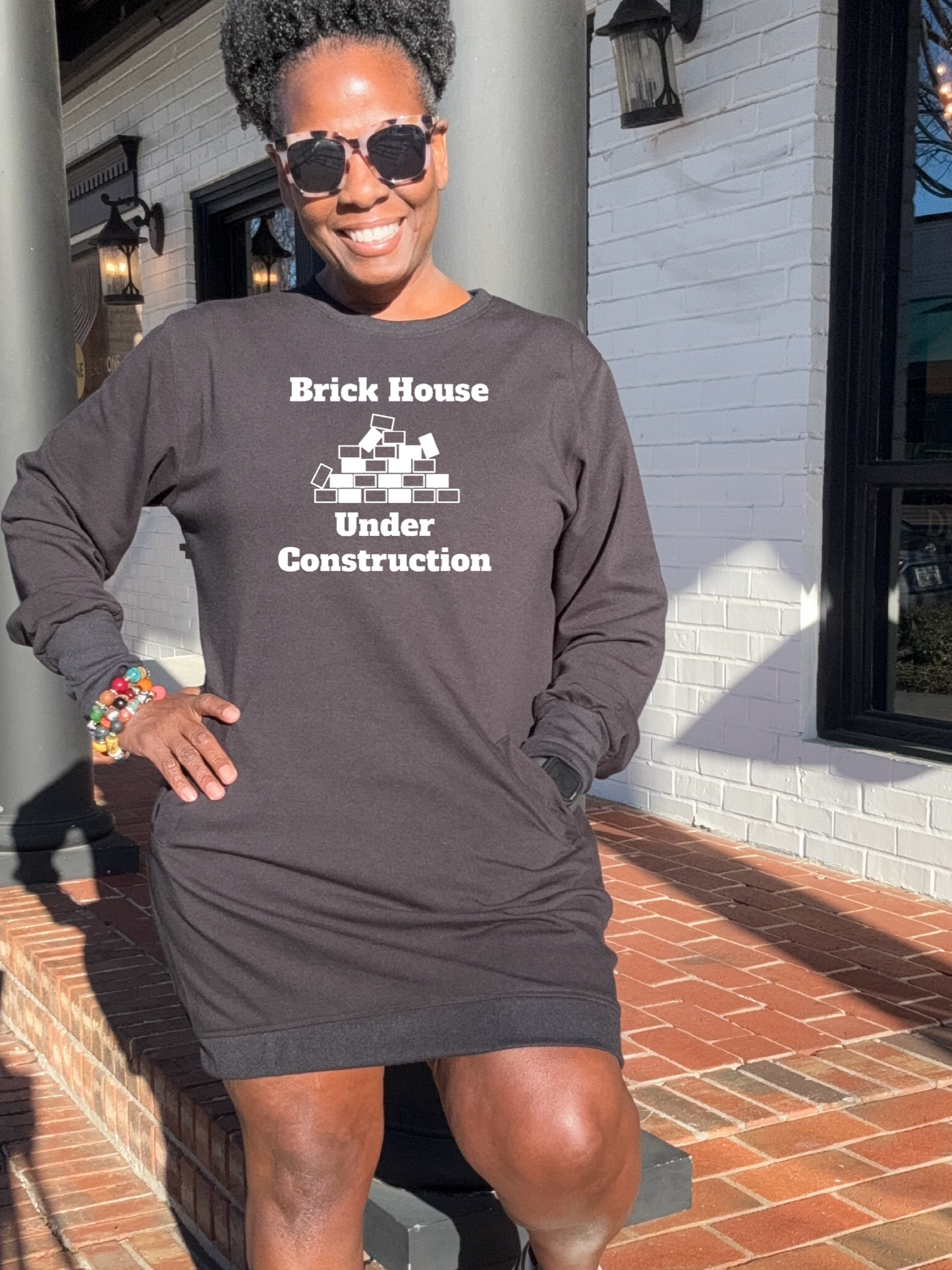 A Black woman standing on a brick pathway wearing a Black sweatshirt dress with the text 'Brick House Under Construction' on it, paired with sunglasses, and gym shoes