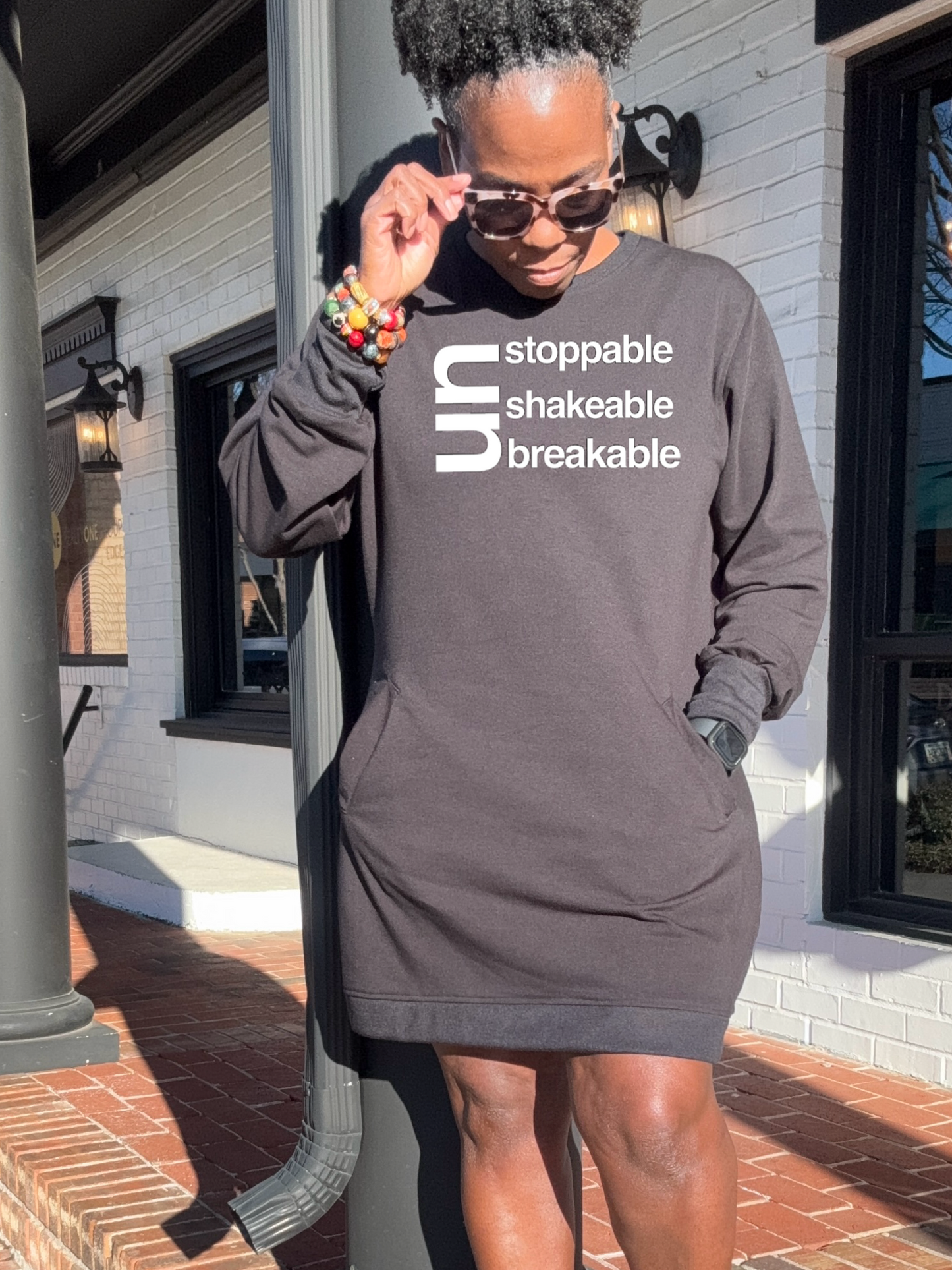 A Black woman standing outside wearing a BLack sweatshirt dress with text 'Unstoppable Unshakable Unbreakable' written across the front. She has accessorized with multiple bracelets and is wearing sunglasses.