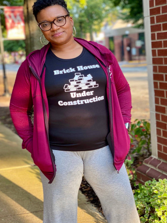 Black girl wearing glasses , gray sweats and a activewear tank top. the tank is black and says Brick house under construction.