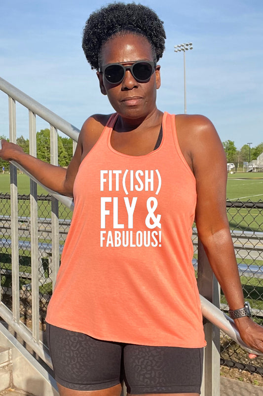 A woman wearing a pink racerback tank top with the text 'FIT(ISH) FLY & FABULOUS' printed on the front, standing near a sports field.