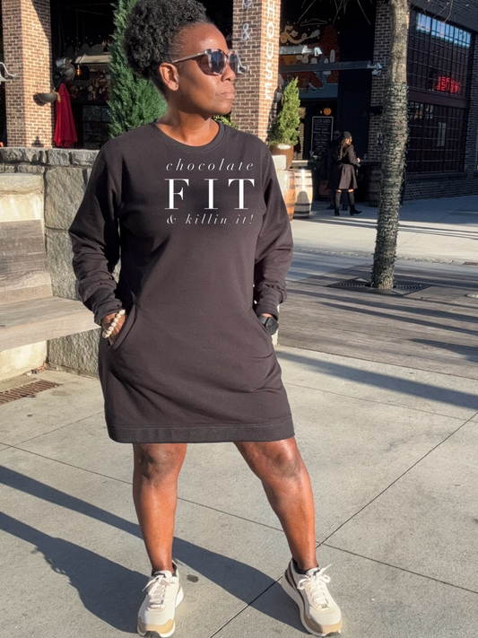 A Black Woman standing on a sidewalk wearing a Black sweatshirt dress with white text 'chocolate fit & killin it', paired with sunglasses and white sneakers.