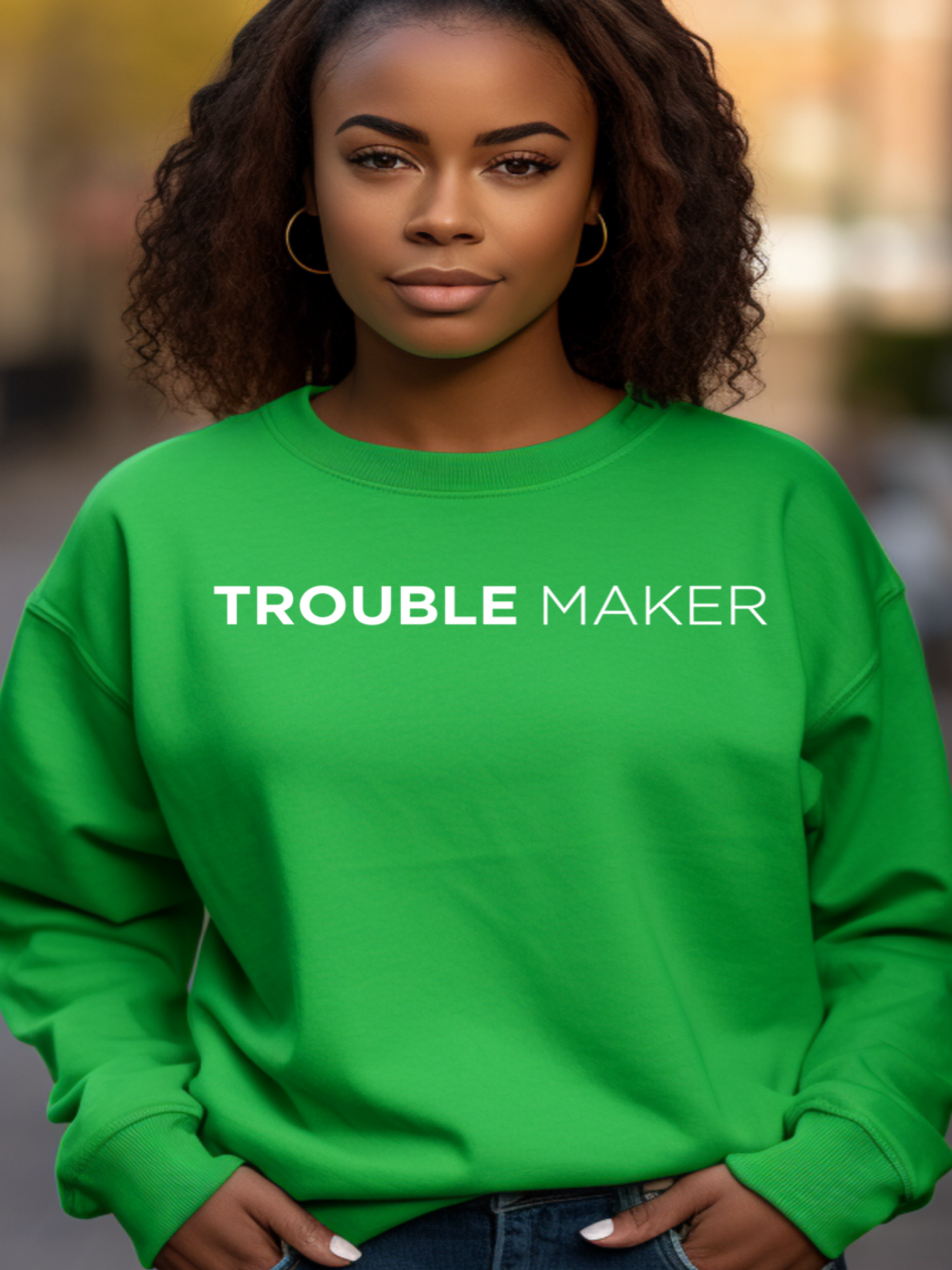 A woman wearing a green crewneck sweatshirt with the text 'TROUBLE MAKER' printed on the front.