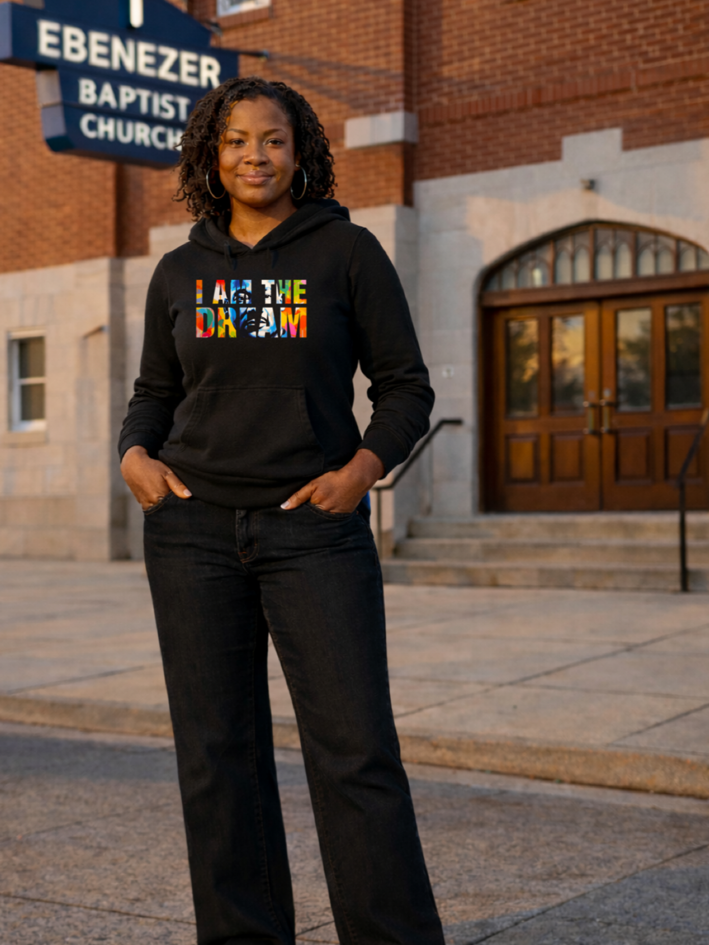 Black lady wearing a black hoodie with text that says I am the dream,  standing front of Ebenezer Baptist Church.