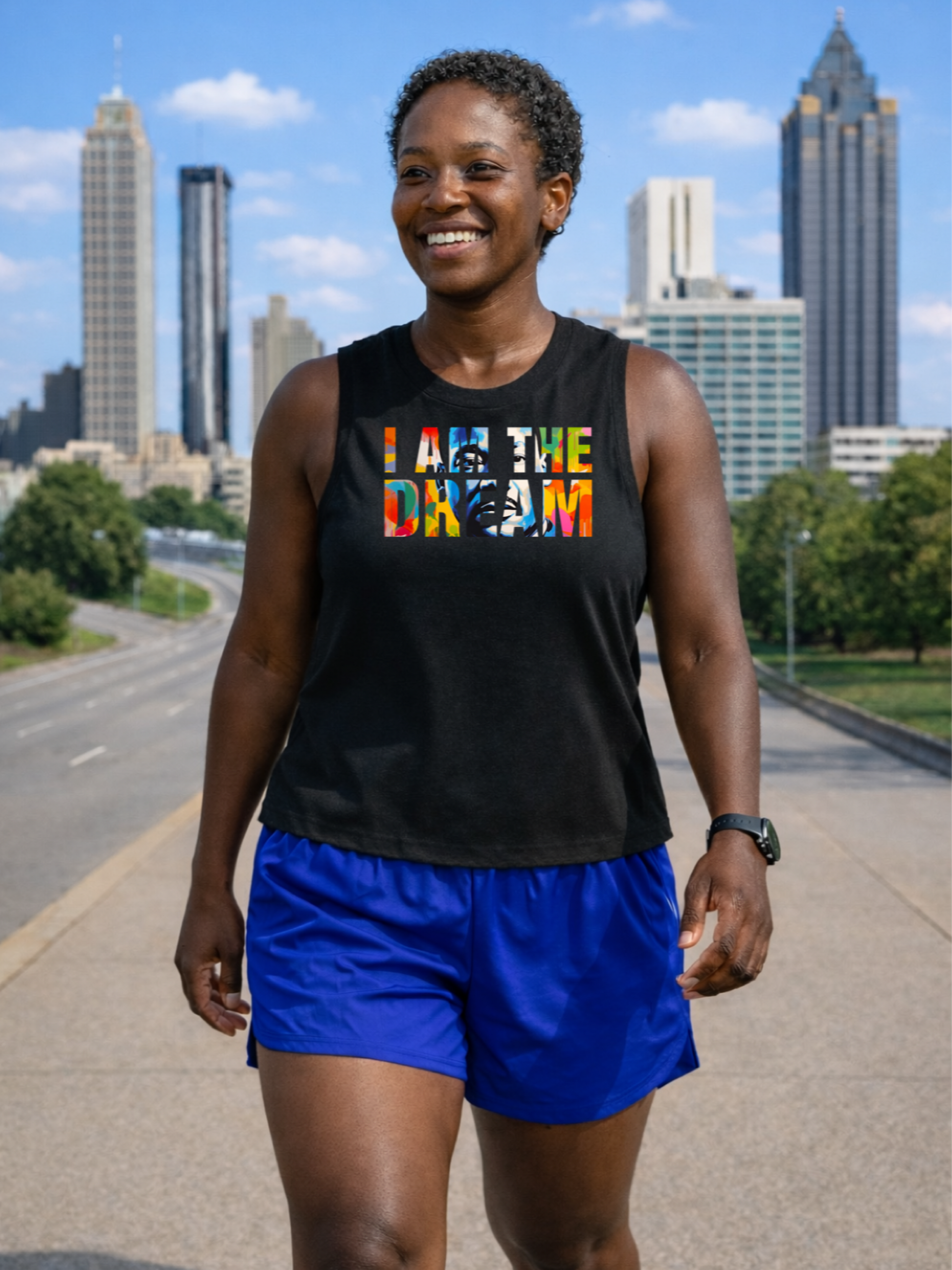 A black runner wearing a black racerback crop tank with colorful text that reads "I am the dream" and blue shorts, standing on a road with a city skyline in the background.