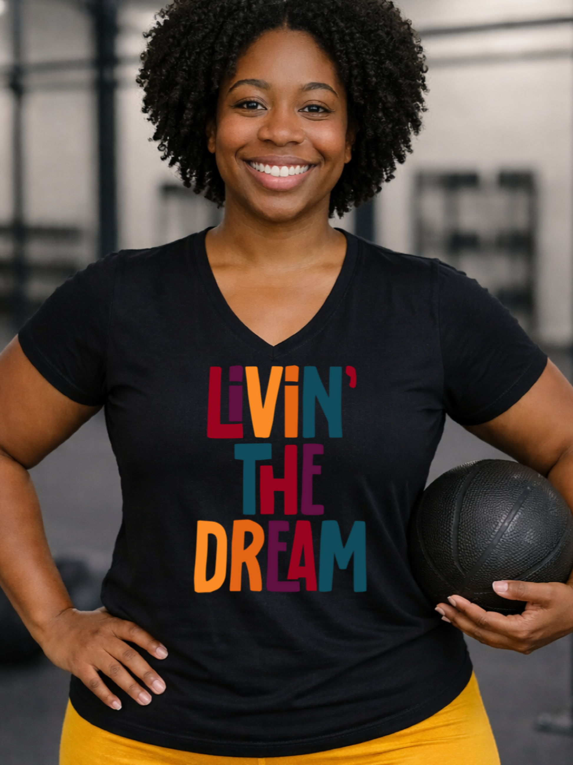 Woman wearing a black t-shirt with 'Livin' The Dream' in colorful letters, holding a medicine ball in a gym setting.