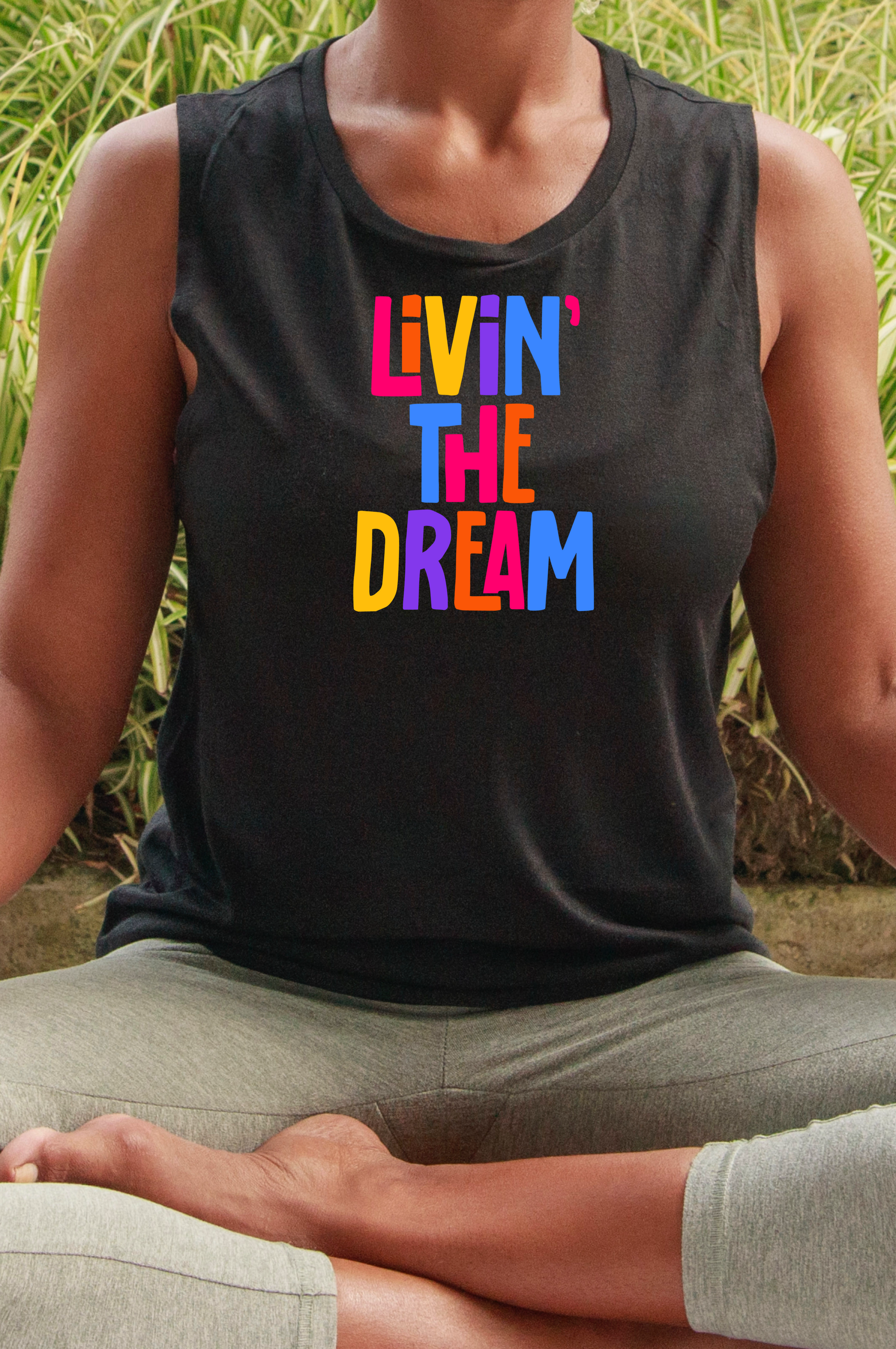 A person wearing a black racerback crop tank top with the text 'LIVIN' THE DREAM' printed in colorful letters,  sitting in a yoga pose 
