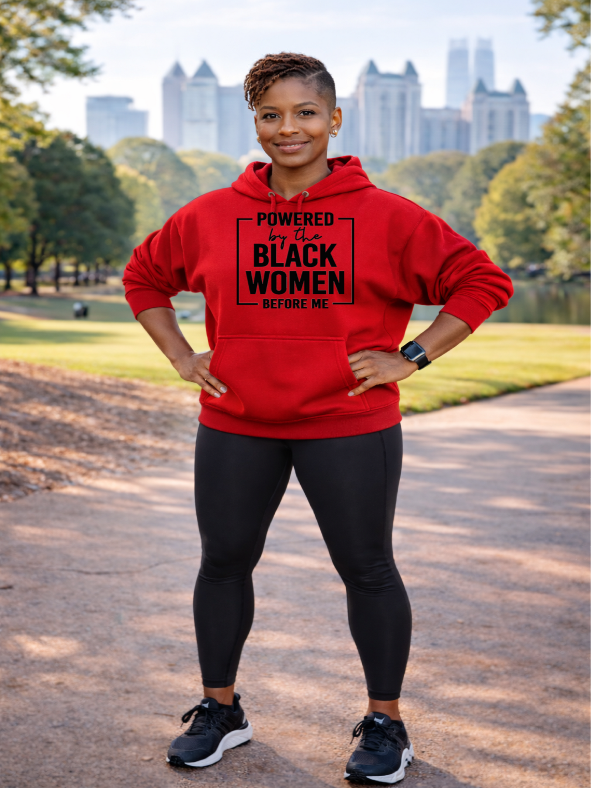 Black lady wearing a red hoodie that says Powered By the Black Women Before  standing in a park with a city skyline in the background