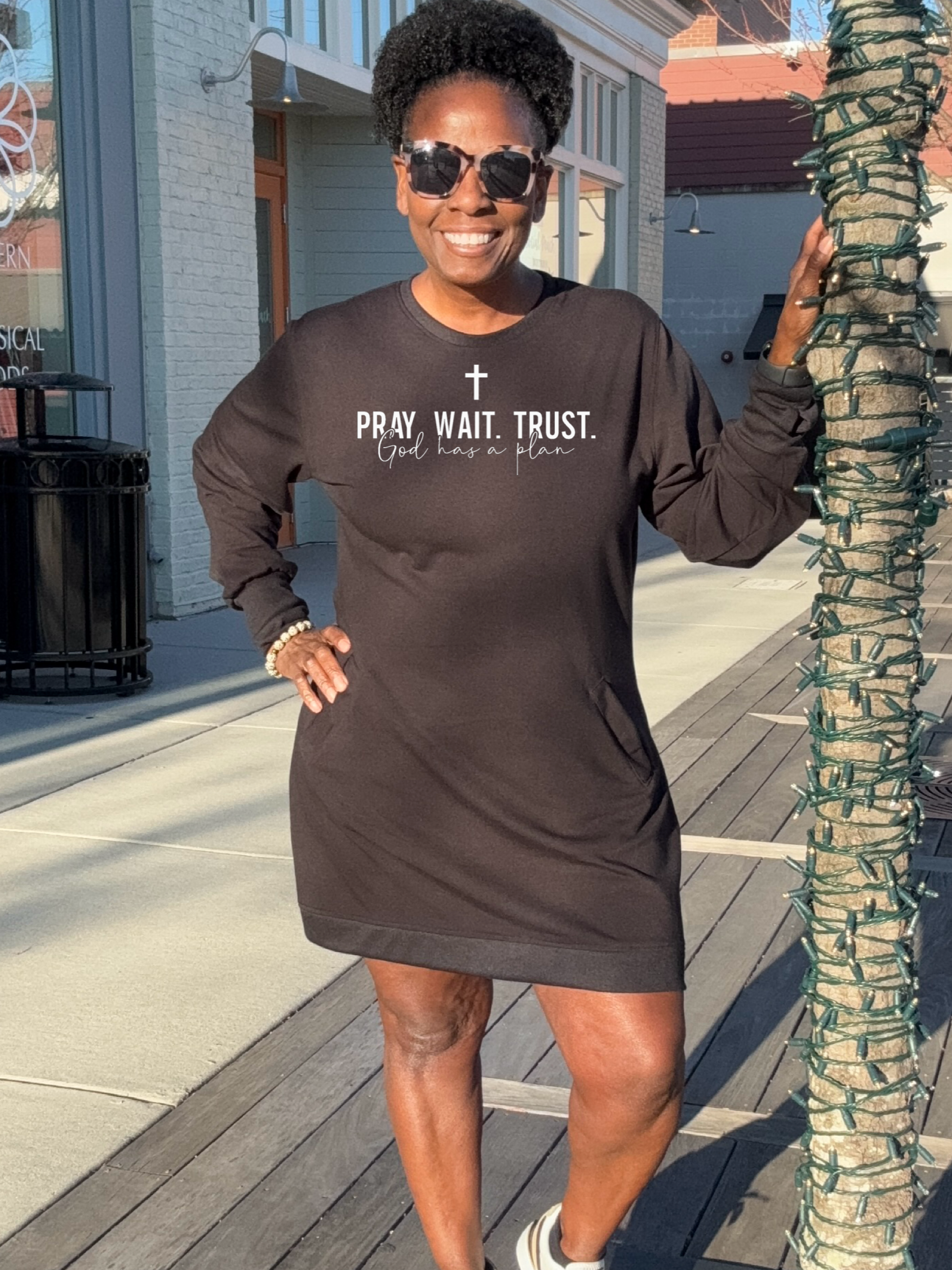 A Black woman wearing a black sweatshirt dress that reads 'Pray, Wait, Trust God Has a Plan' stands outdoors.