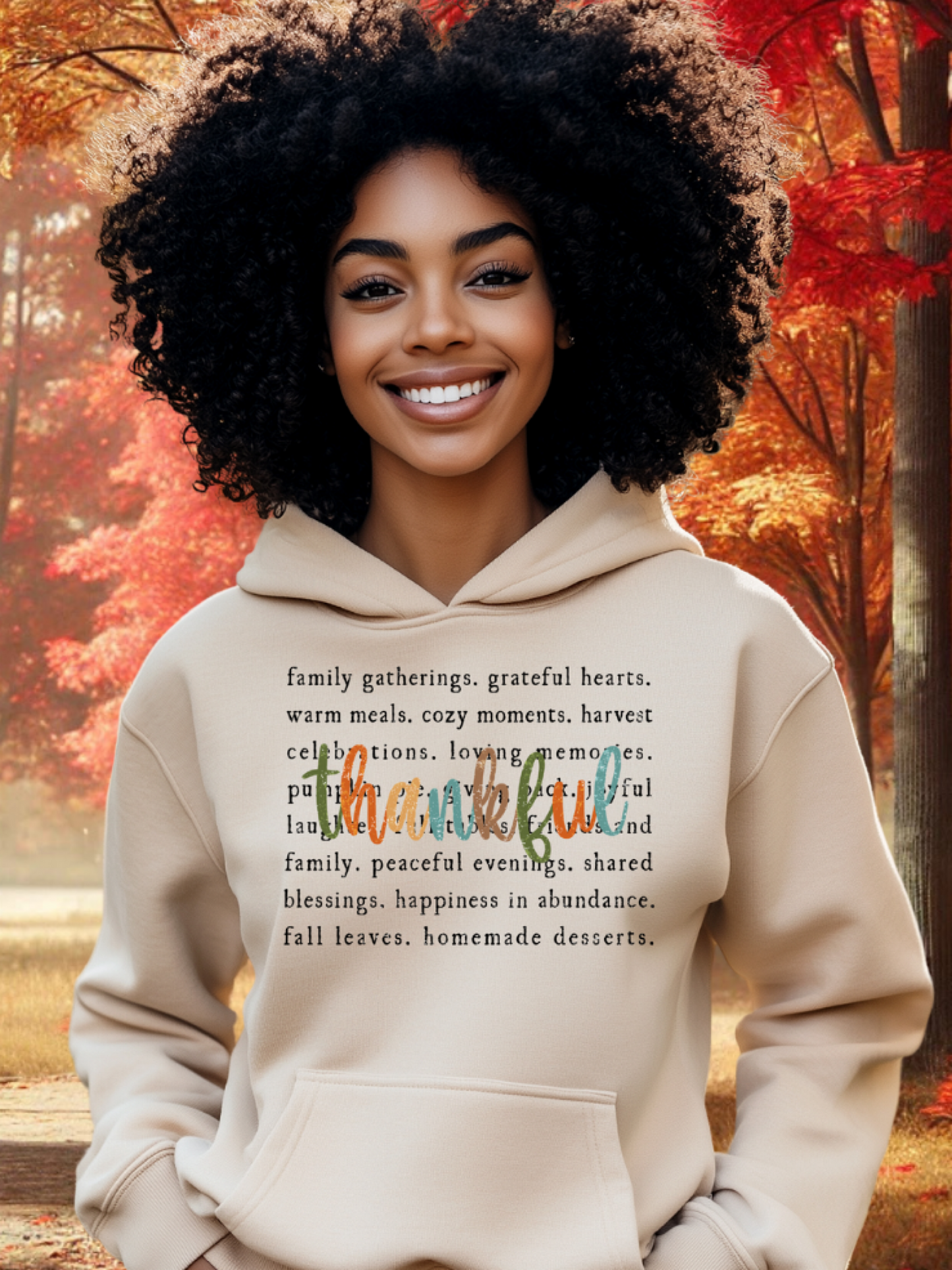 A Black woman smiling and wearing a beige hoodie with a text design that includes the words 'thankful', 'warm', 'cozy', and other positive adjectives, standing in a park with autumn trees in the background.