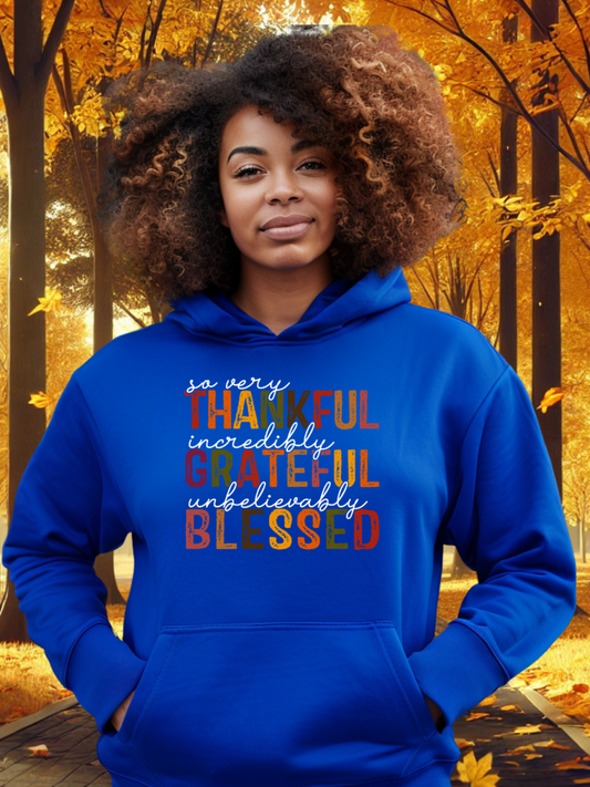 A woman wearing a blue hoodie with thankful, grateful, and Blessed text on it, standing in a park with autumn leaves around.