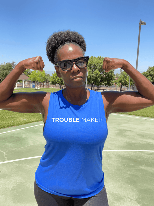 Black woman with biceps pose wearing a royal blue muscle tank that says Trouble Maker 